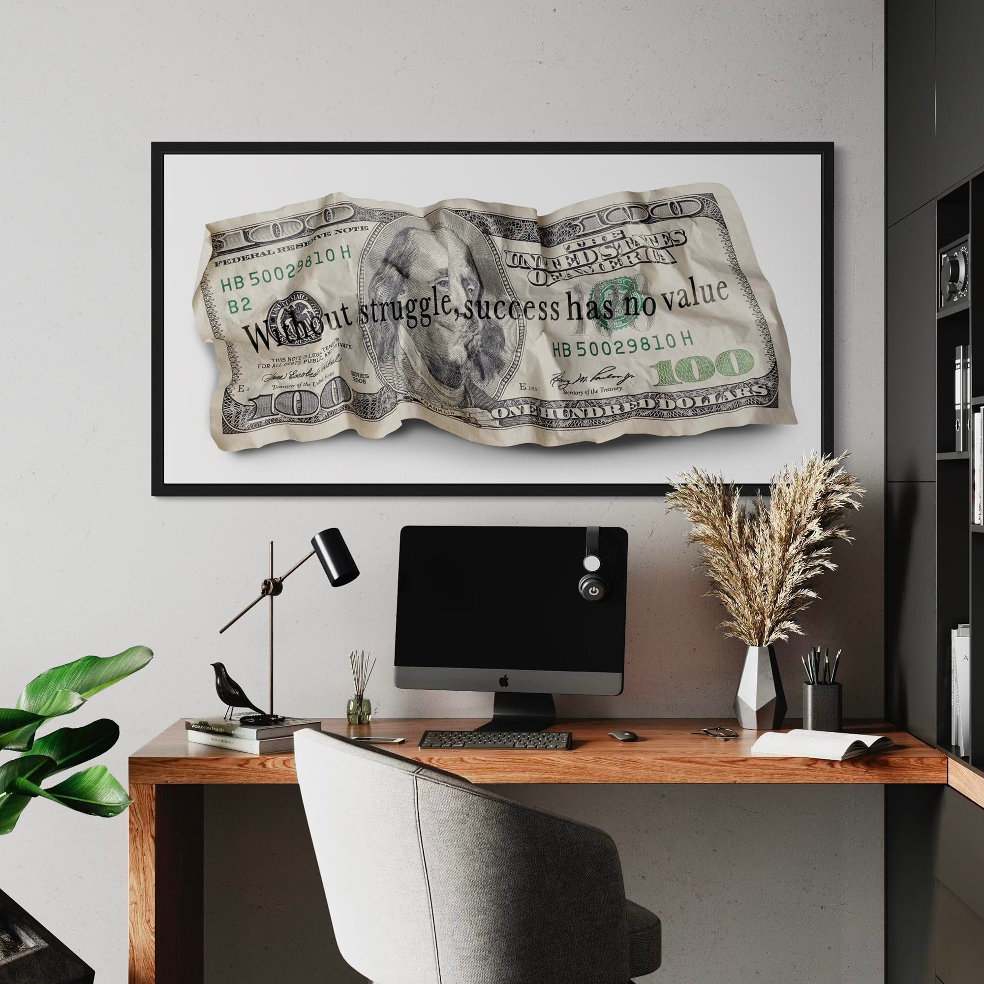 Framed artwork of a crumpled dollar bill with motivational text above a desk setup.